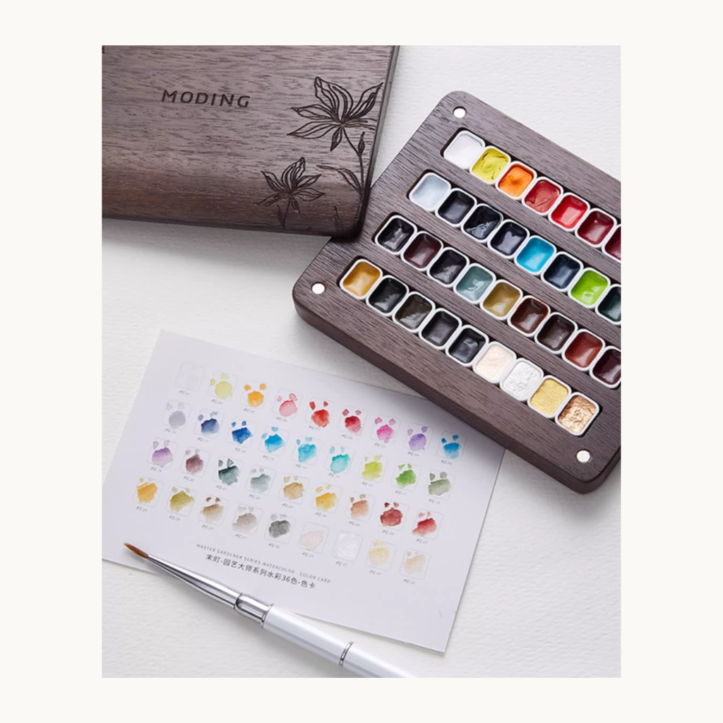 Hand holding brush while mixing MODING 36-color watercolors on a wooden mixing board, with full swatch sheet displayed.