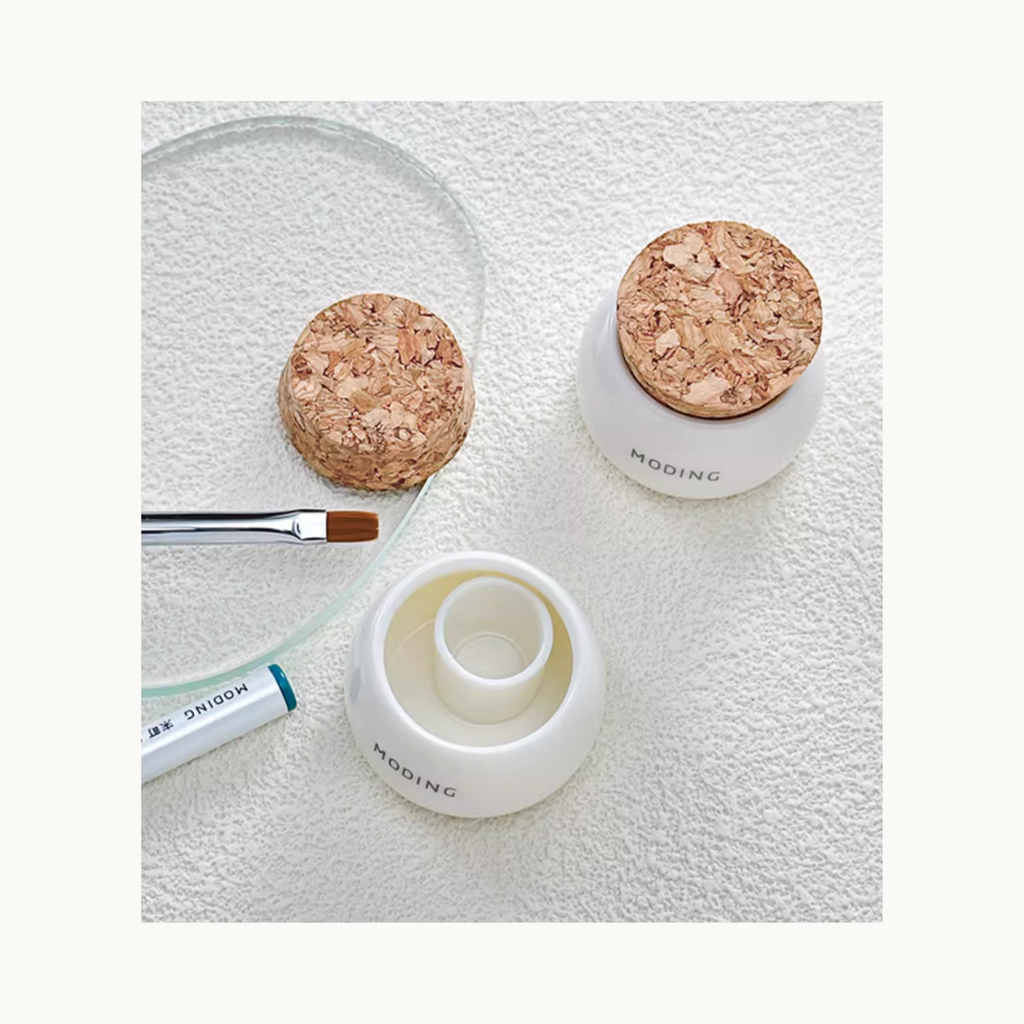 EN: Top-view display of Moding ceramic brush cleaner cup with cork lid and nail brush on a soft textured background.