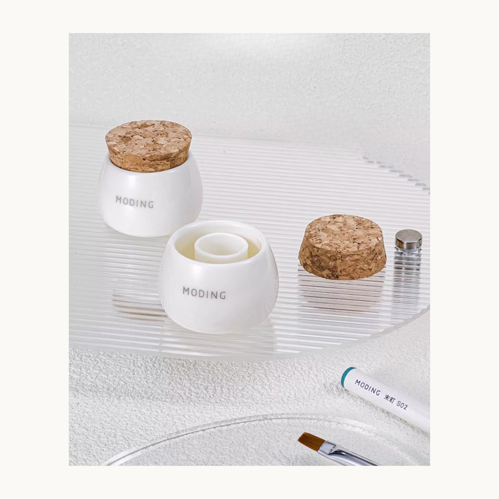 EN: Ceramic dual-chamber brush cleaner cup with cork lid, displayed on a textured surface with nail tools.