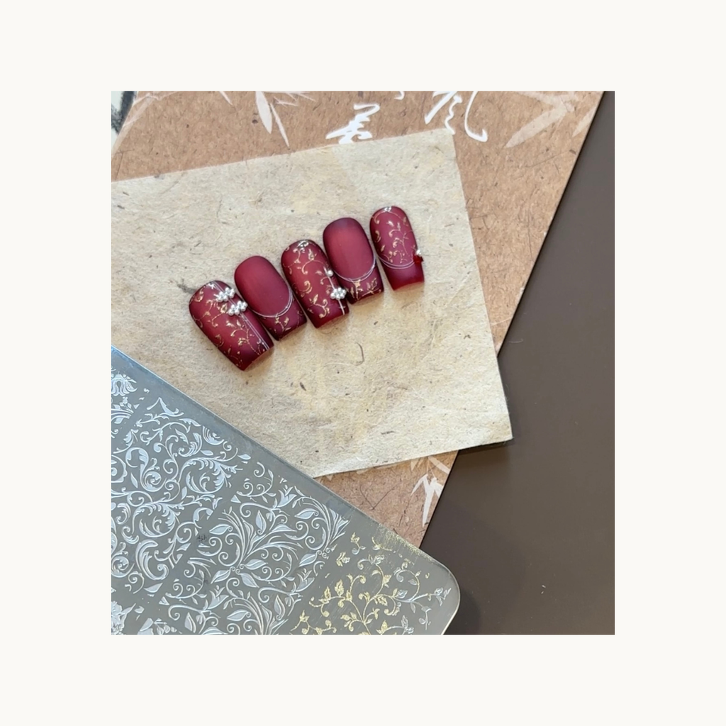 Pearl-accented burgundy nails featuring fine gold stamped patterns made with QingYou gold transfer paste.